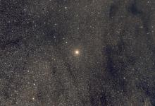 A bright red star on a background of dense faint stars and some dark dust sweeps.